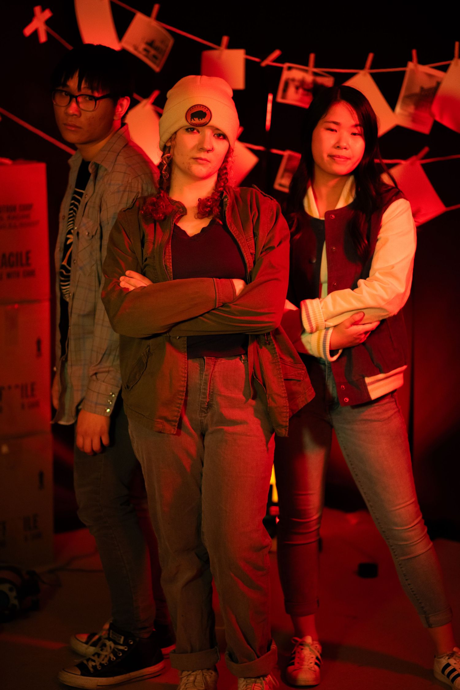 Three young people posing confidently under hanging photos on a dramatic red-lit set.