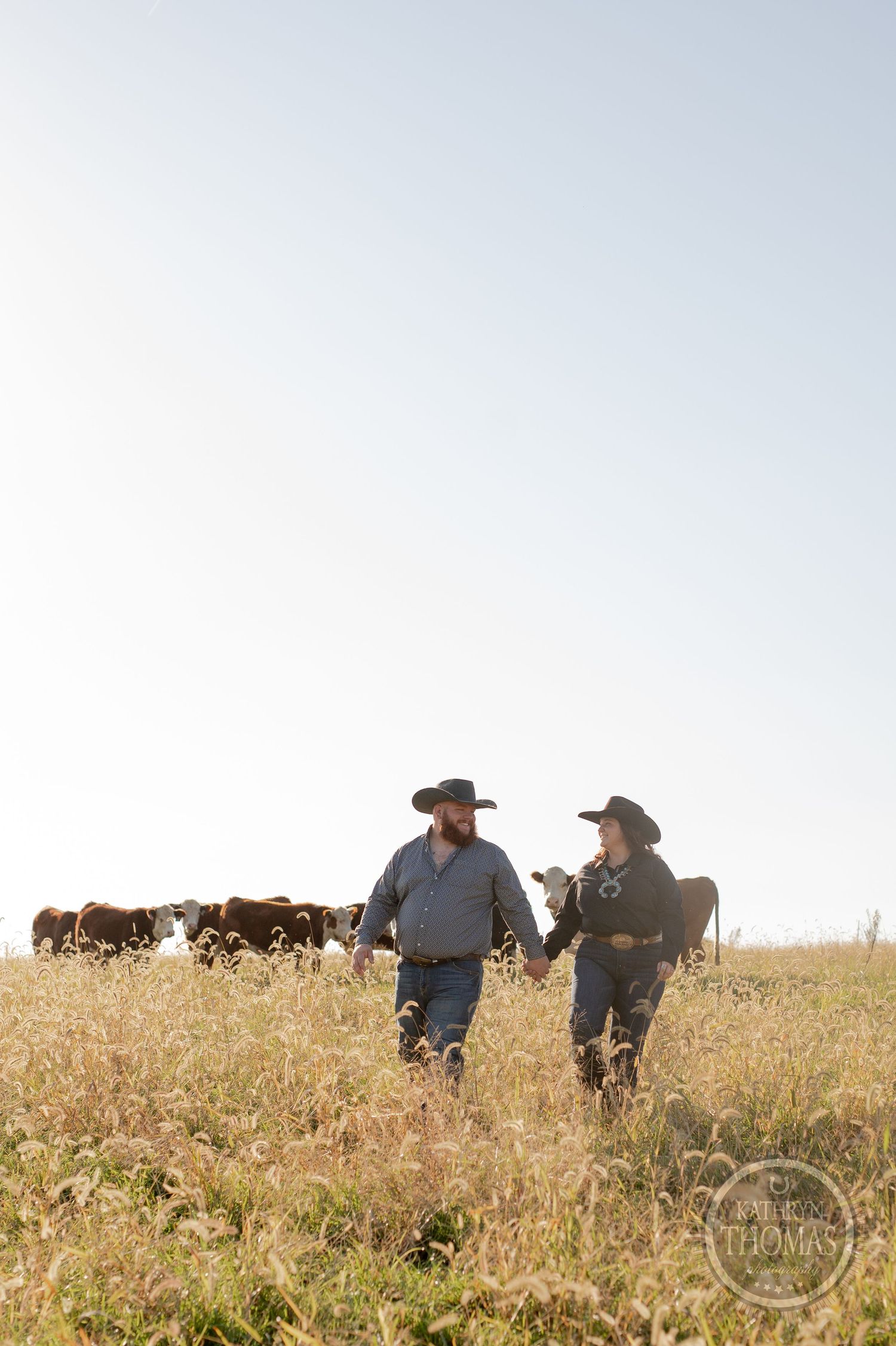 Two ranchers in cowboy hats walking through a golden field with cattle grazing in the background.
