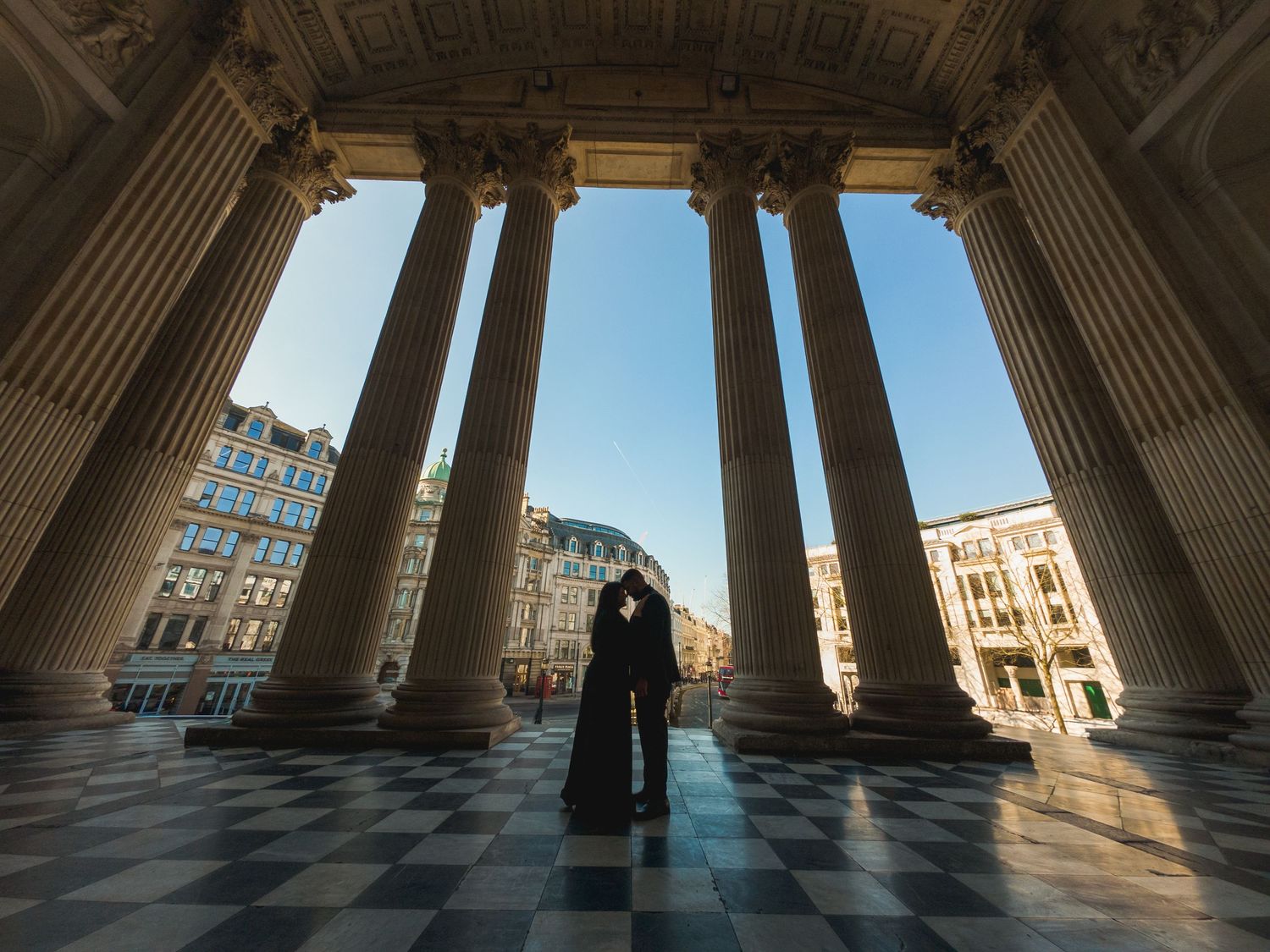 A silhouetted figure stands between classical columns at St. Paul's Cathedral in London, with checkered floor and city view beyond.