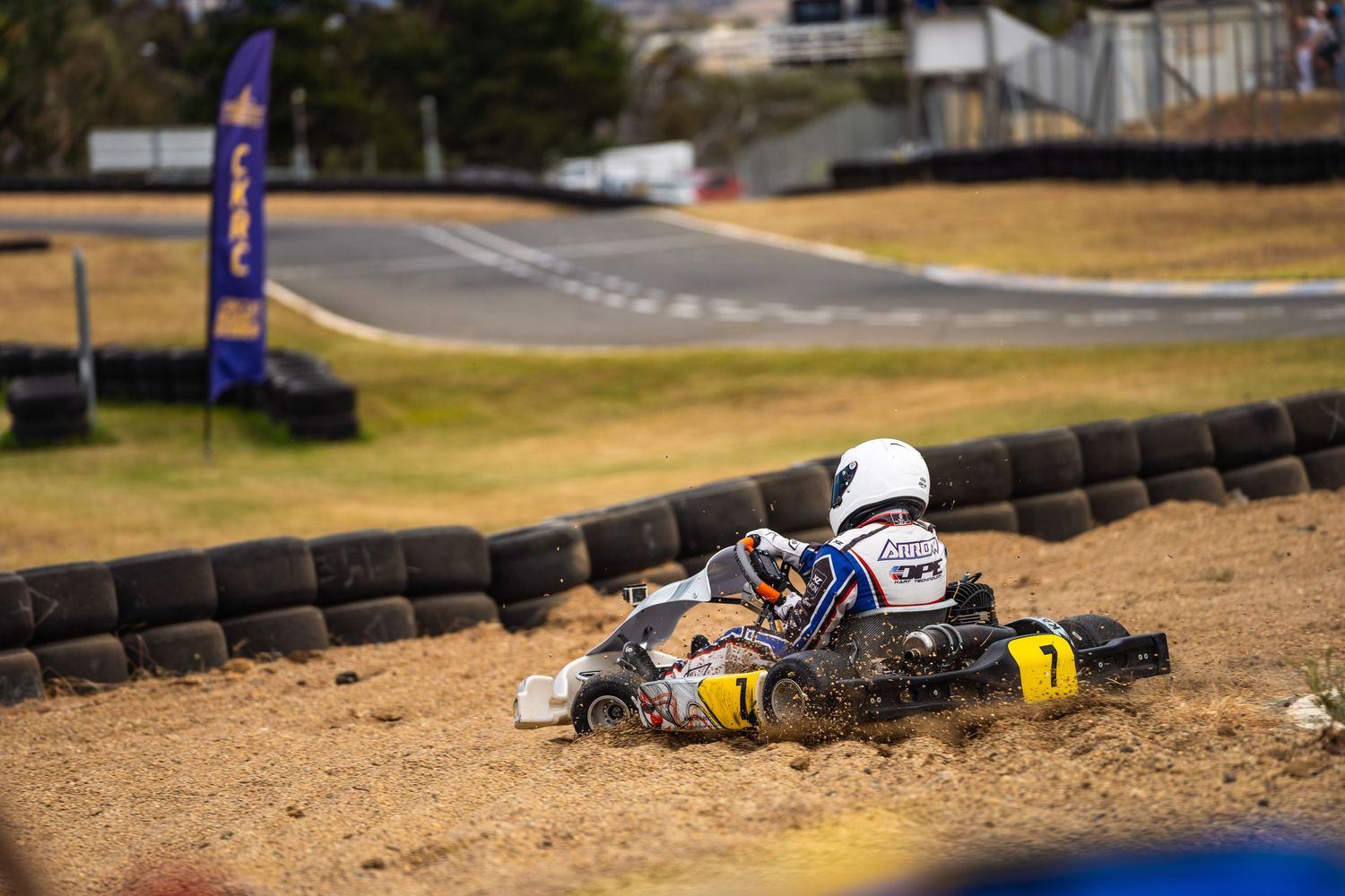 A go-kart racer navigates a turn on a dirt track with tire barriers and a race flag visible in the background.