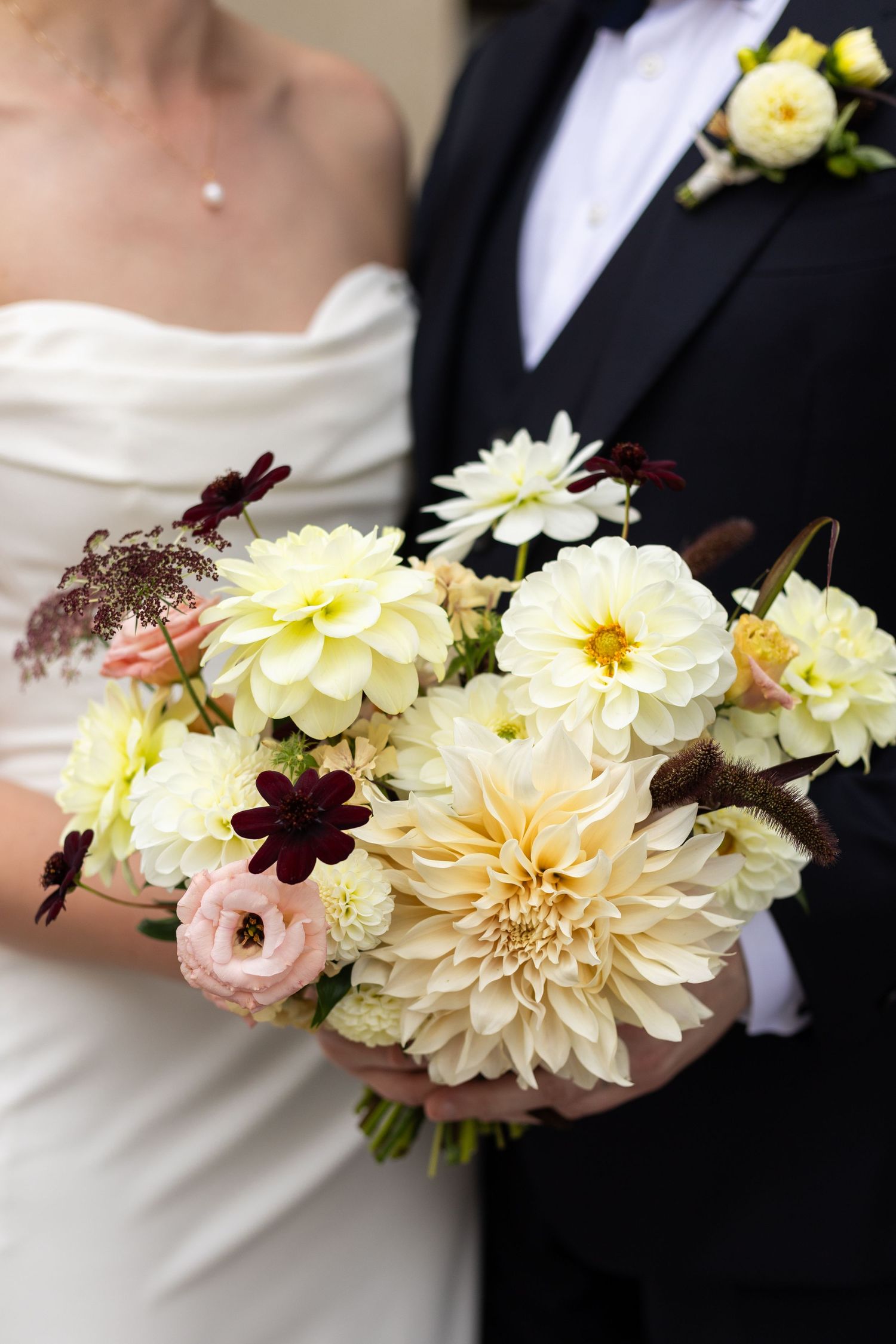 An elegant wedding bouquet with cream dahlias and burgundy accents held between bride and groom.