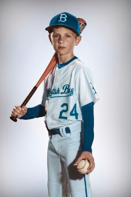 Young baseball player in white and blue uniform holds bat and ball while posing in classic portrait style.