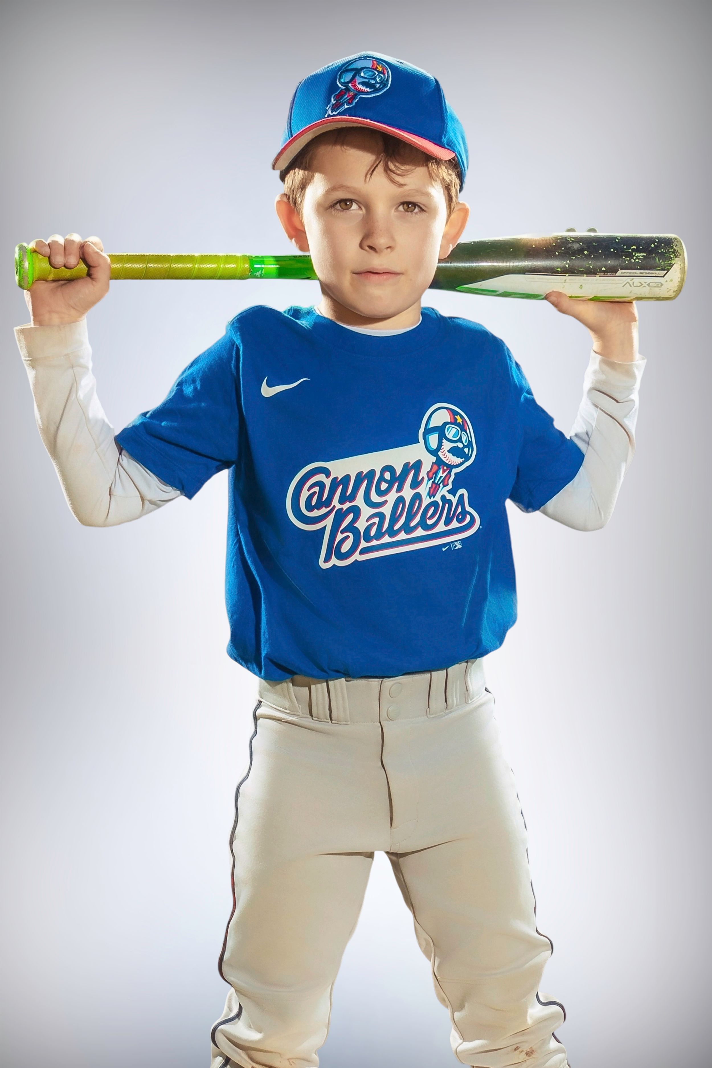 Young baseball player poses in blue team uniform with baseball bat against light background.