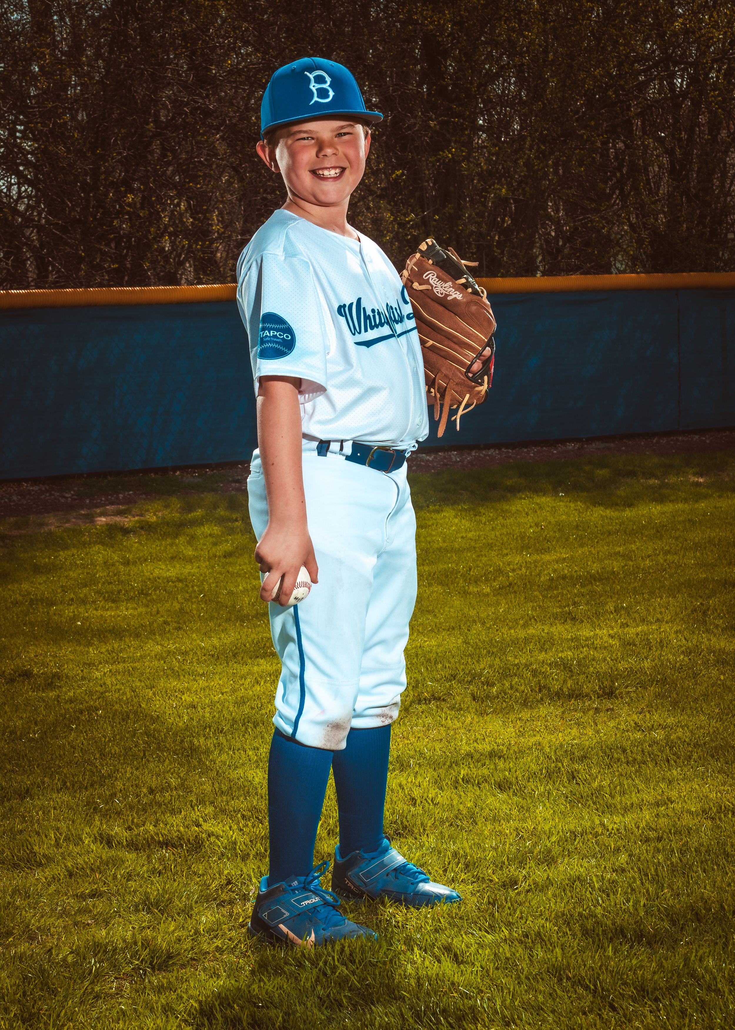 Youth baseball player wearing light blue uniform and cap stands with glove on baseball field at practice session.