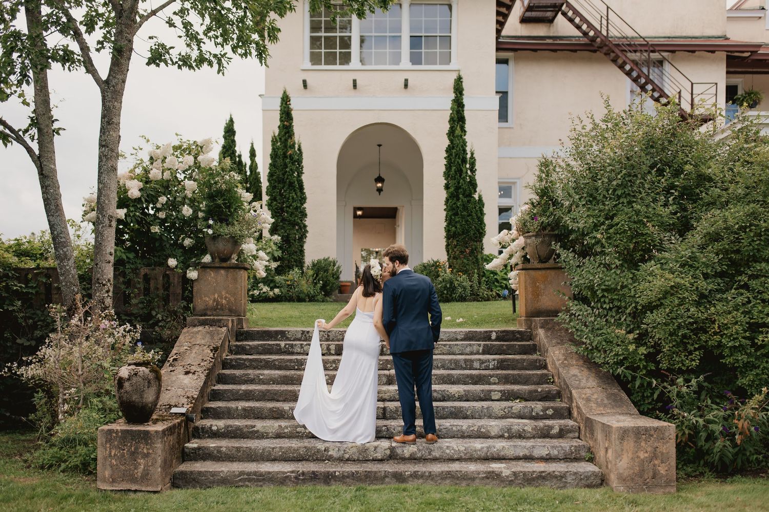 A couple stands on grand stone steps leading to an elegant Mediterranean-style villa.
