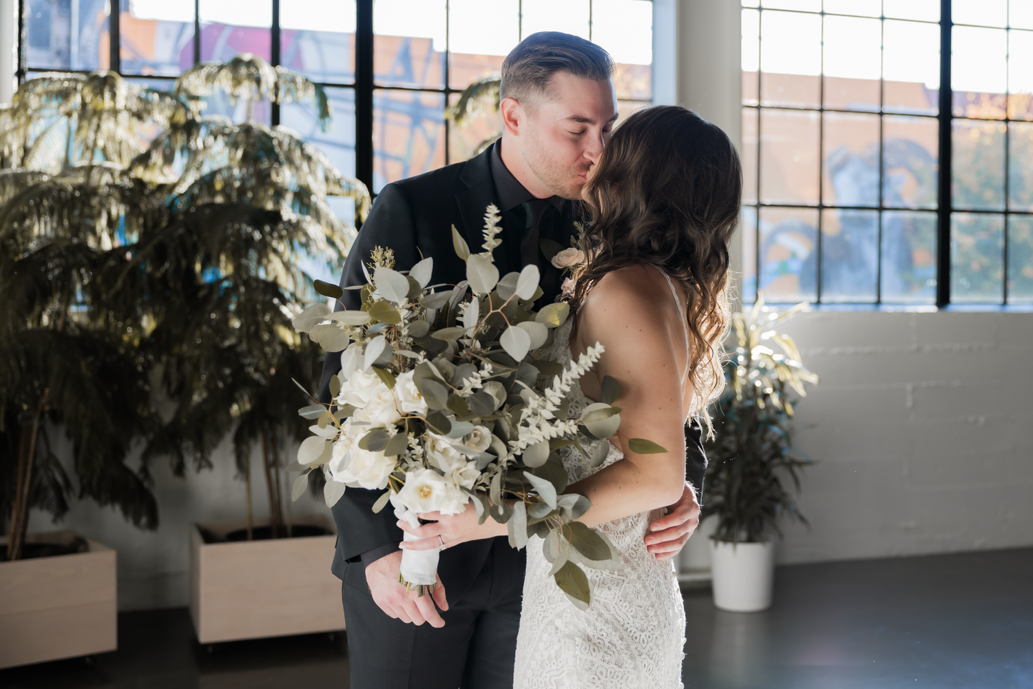 A romantic wedding moment between a couple embracing near large windows, surrounded by elegant floral arrangements.