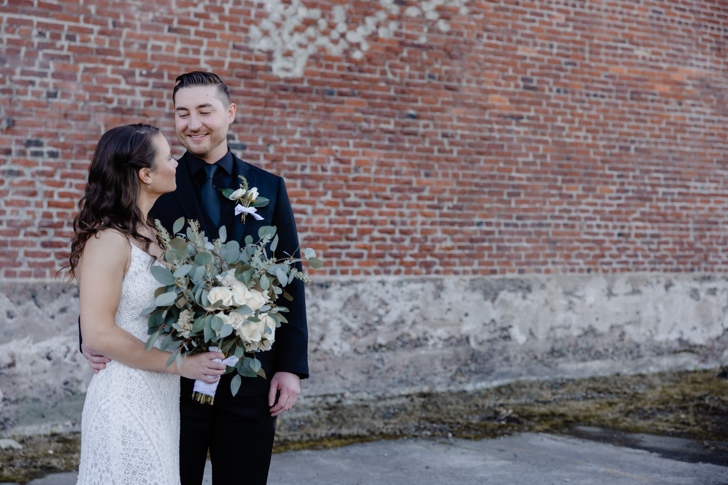 Newlywed couple shares romantic moments against rustic brick wall backdrop during wedding photo session.