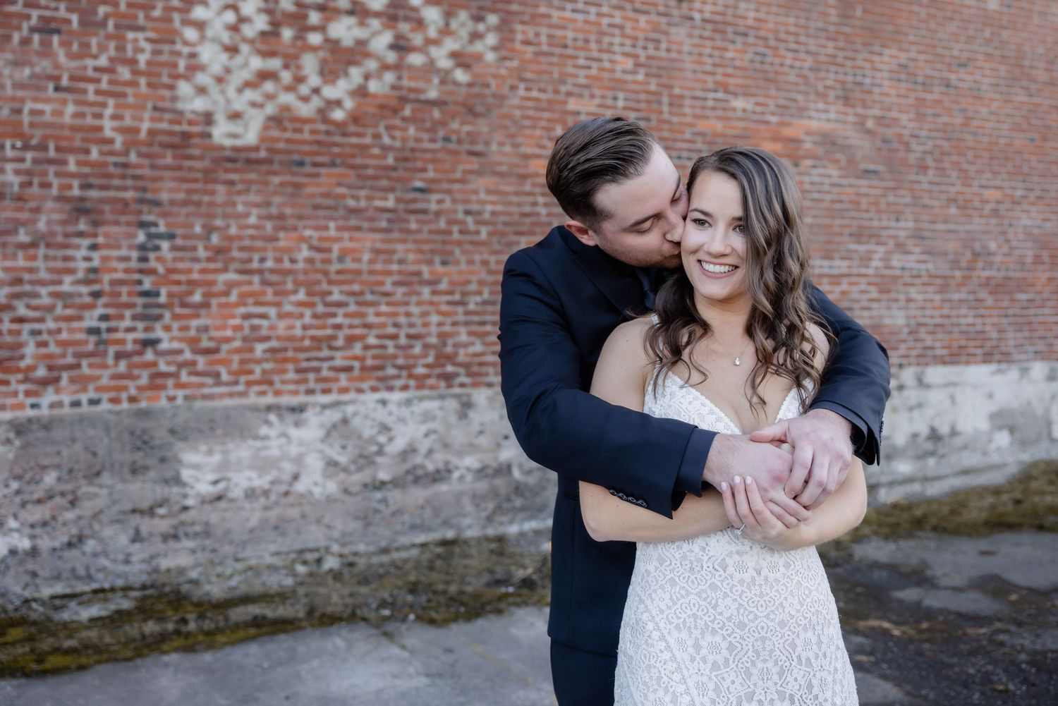 A couple in formal attire shares romantic moments against a rustic brick wall backdrop.