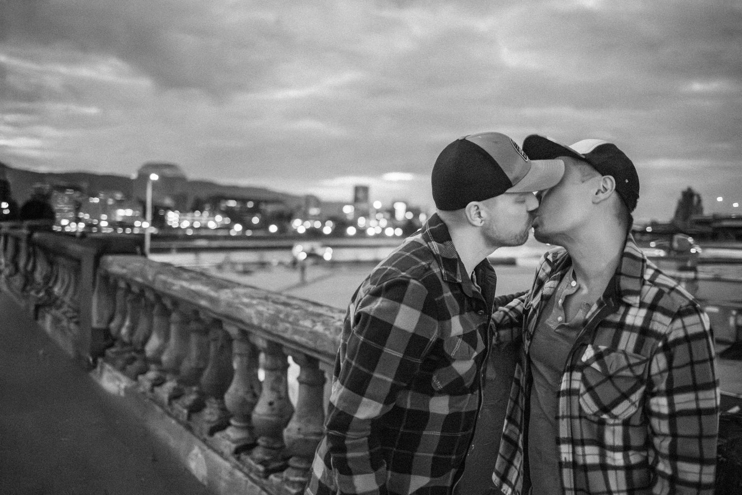 A couple in matching plaid shirts share a tender moment along a stone balustrade with a cityscape in the background.
