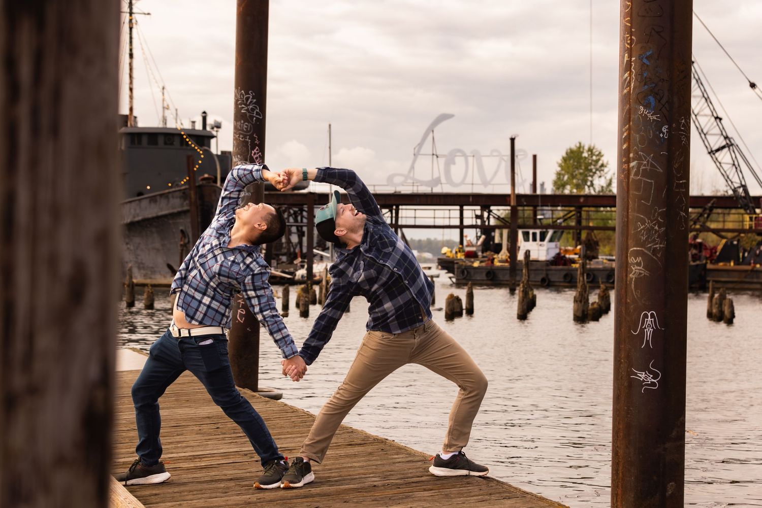Two people in casual wear playfully dance together on a wooden dock by the water with a bridge visible in the background.