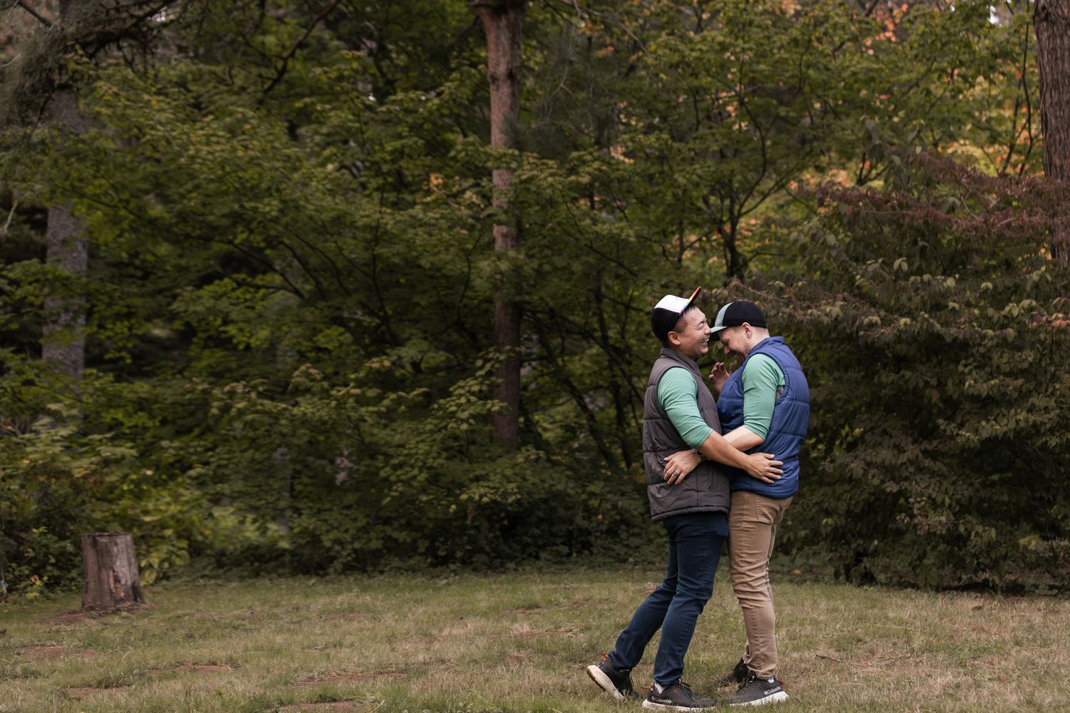 A couple in casual attire performs a romantic dance dip move outdoors in a wooded park setting.
