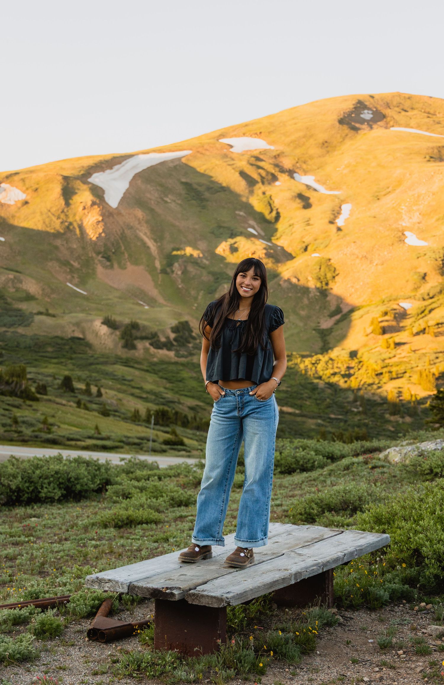Person stands on a wooden bench at sunset in the mountains with golden light illuminating the alpine landscape.