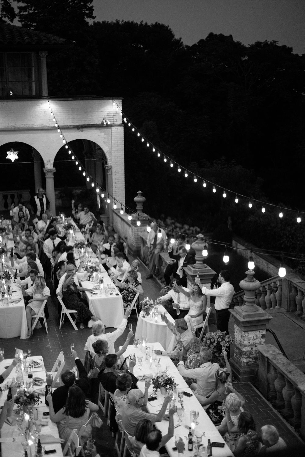 Black and white aerial view of an elegant outdoor wedding reception at dusk with string lights and guests dining on a terrace.
