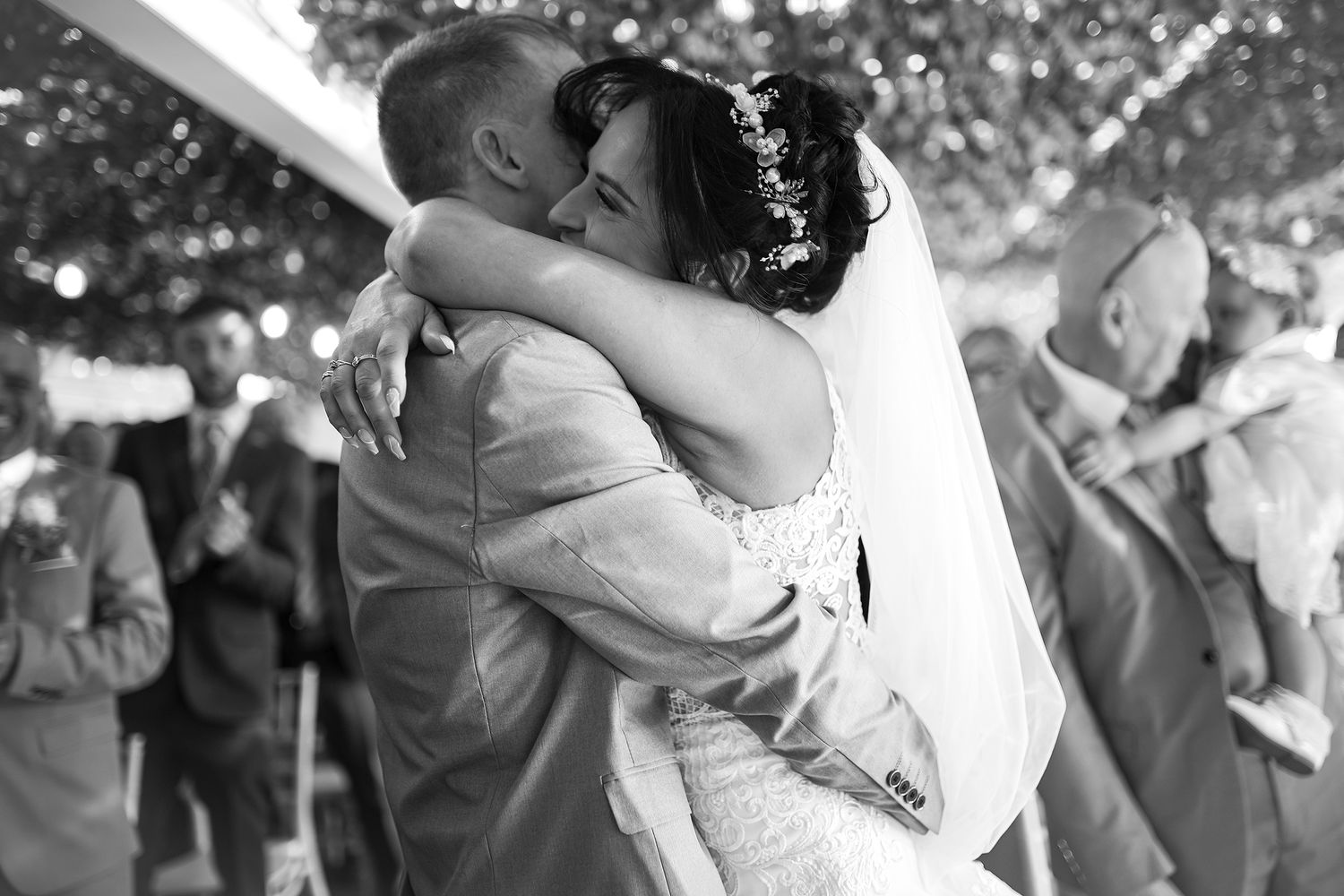 Black and white photo of bride and groom sharing an emotional first dance embrace surrounded by wedding guests.