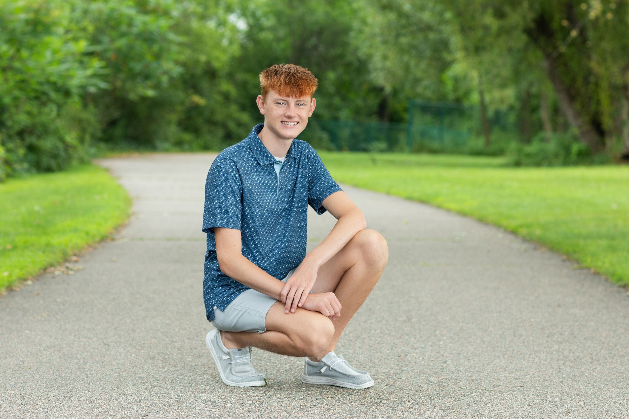 Person in blue polo shirt and khaki shorts crouches on a paved path in a lush green park setting.