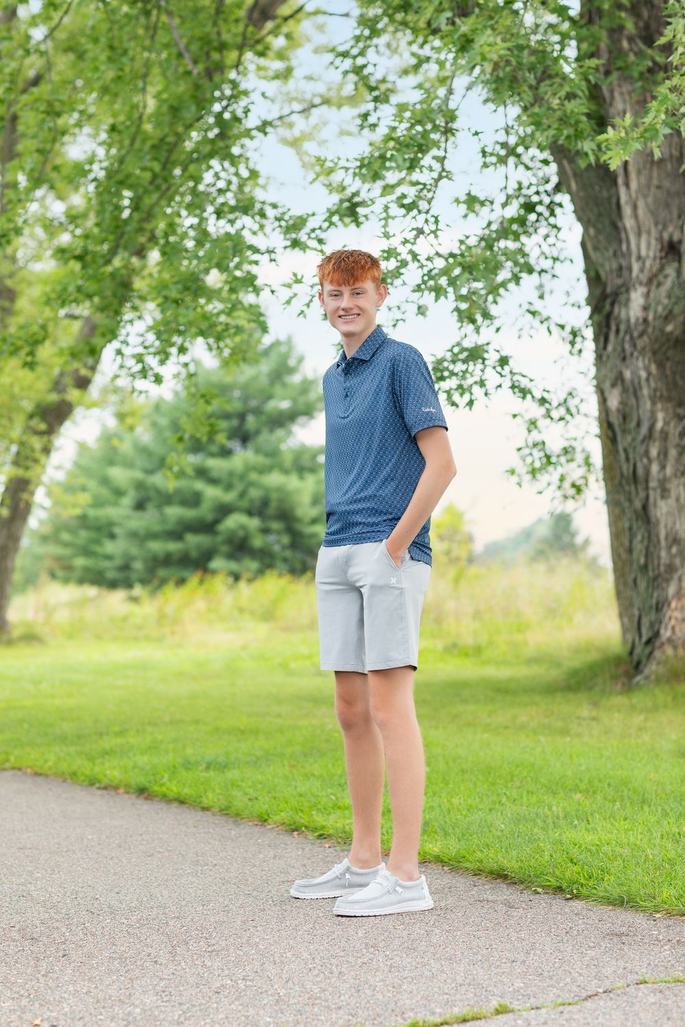 Person in blue shirt and khaki shorts standing on a path surrounded by green trees and grass.
