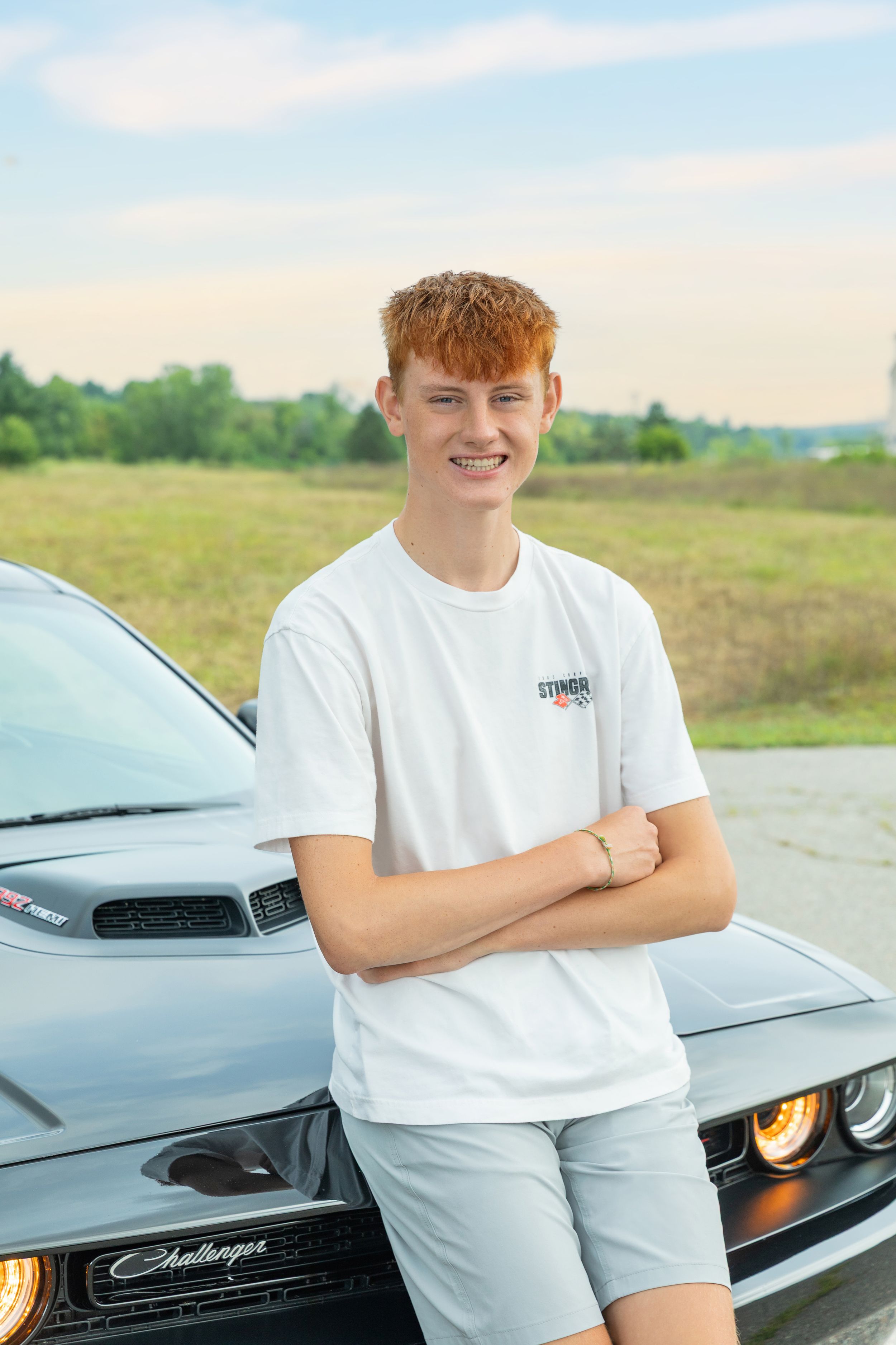 Teenager in white t-shirt stands smiling with arms crossed in front of black Dodge Challenger in sunny field.