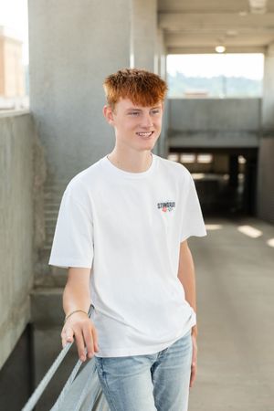 Young person in white t-shirt poses casually against concrete wall in parking structure with natural lighting.
