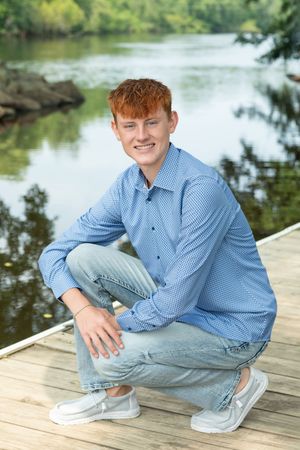Person in blue button-down shirt sitting on wooden dock by peaceful lake.