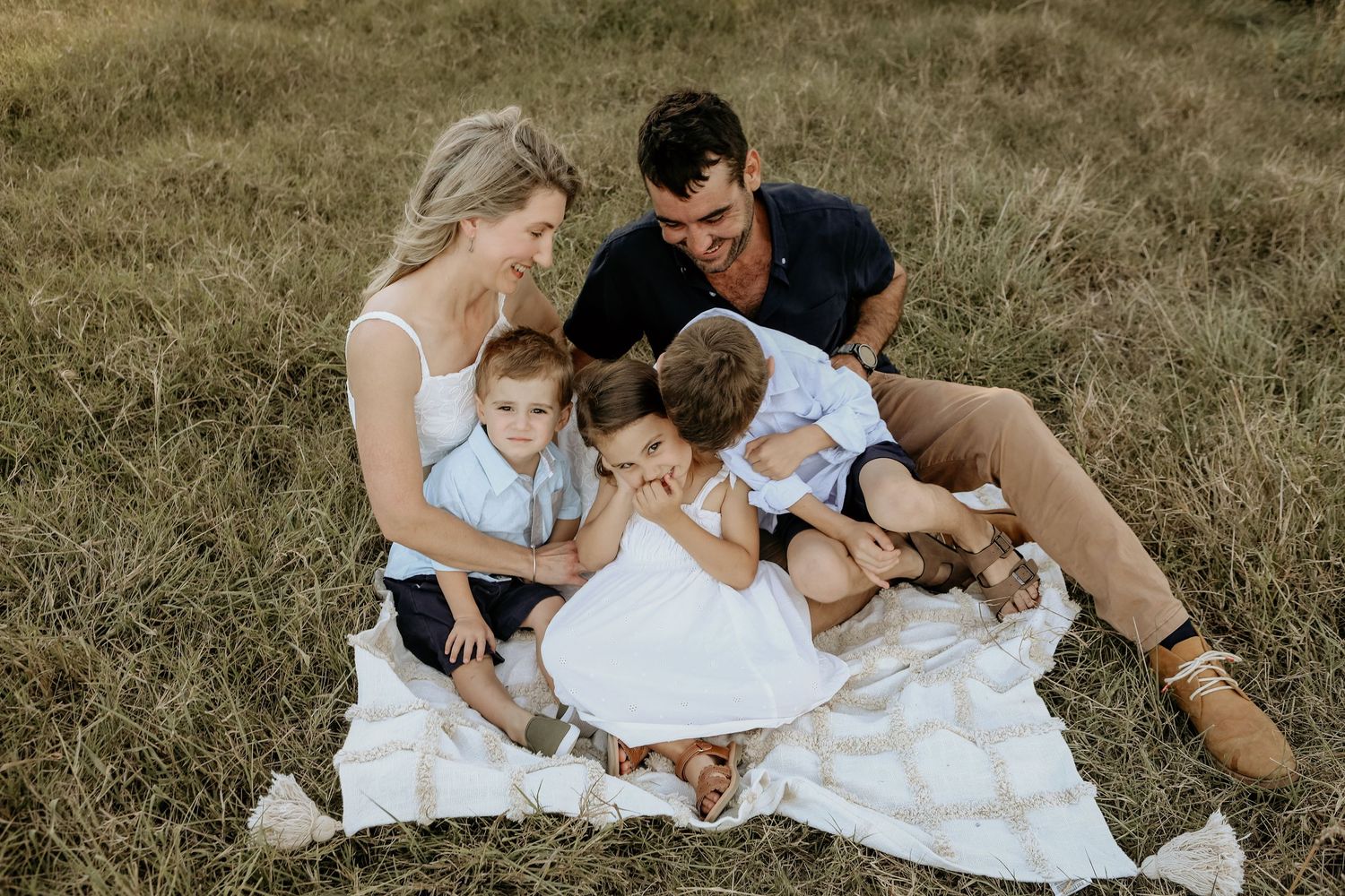 Family wearing white outfits cuddle together on grassy hillside during golden hour photo session.