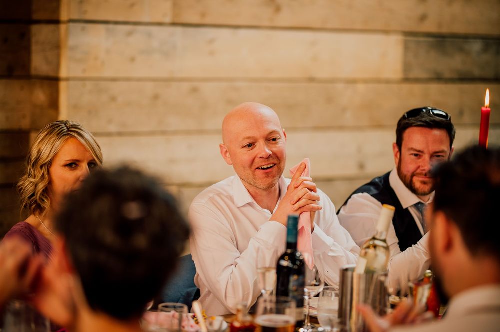 Wedding guests laugh and enjoy themselves during dinner at a rustic reception venue with wooden walls and candlelight.