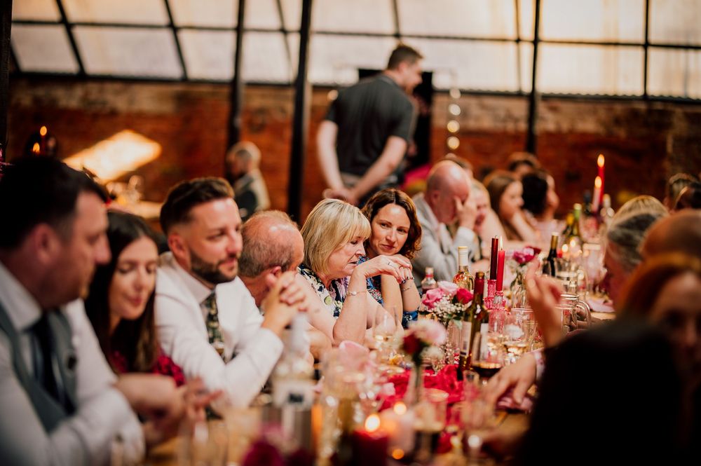 Wedding reception guests seated at long tables with pink floral centerpieces in a warmly lit rustic venue.