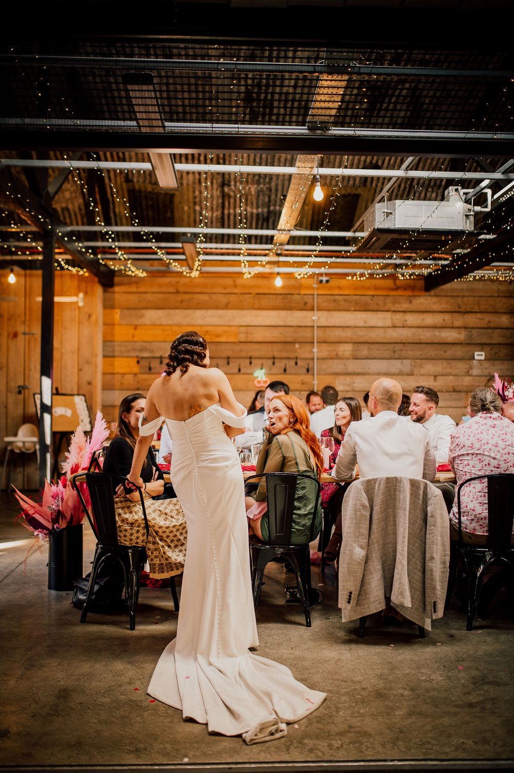A wedding reception in an industrial venue with exposed ceiling and guests seated at long tables.