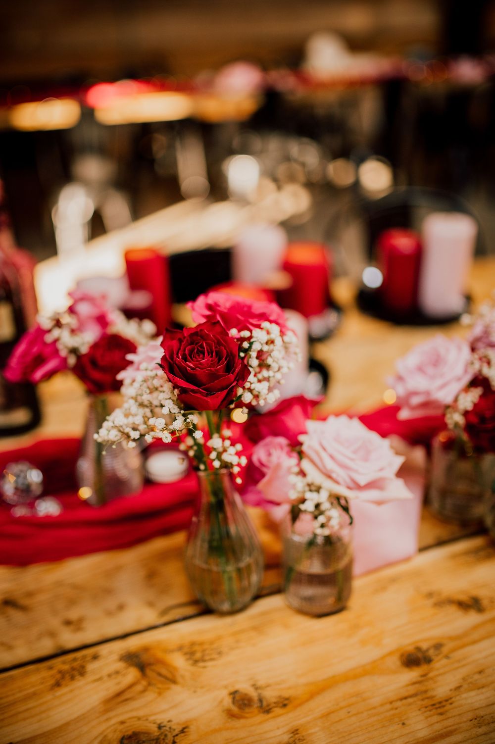 Red and pink roses with baby's breath in mason jars create an elegant rustic table centerpiece on a wooden surface.