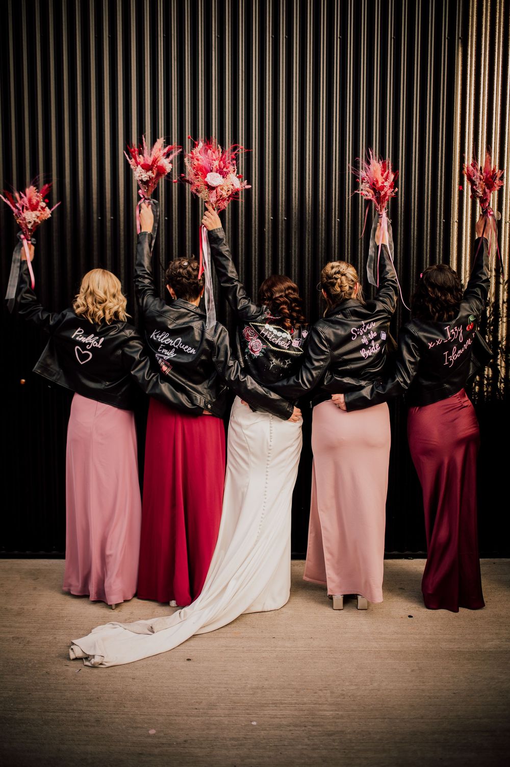 Bridesmaids in pink and burgundy dresses holding pink bouquets with their backs turned to the camera.