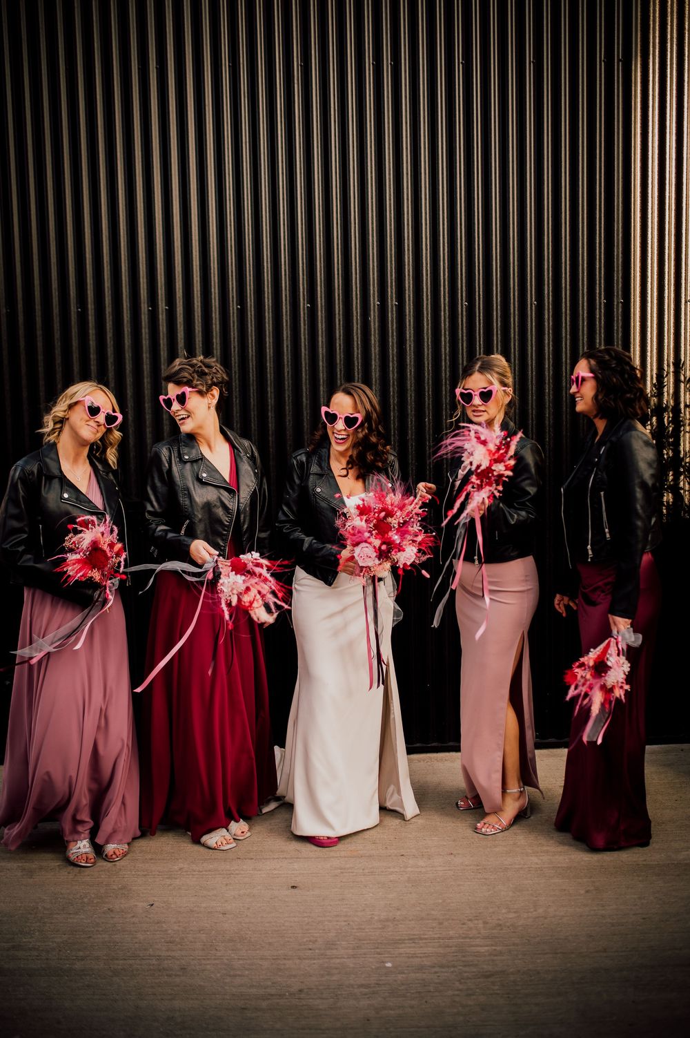 Wedding party in shades of burgundy and pink dresses pose in a line against a dark wall with festive pom poms.