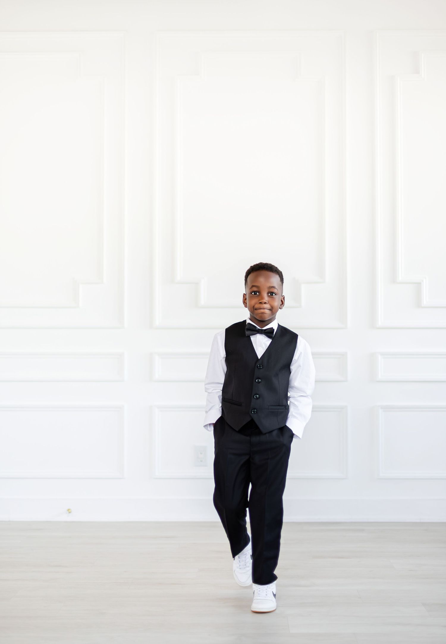 Young child in formal black tuxedo and bow tie stands against white wall in elegant white sneakers.
