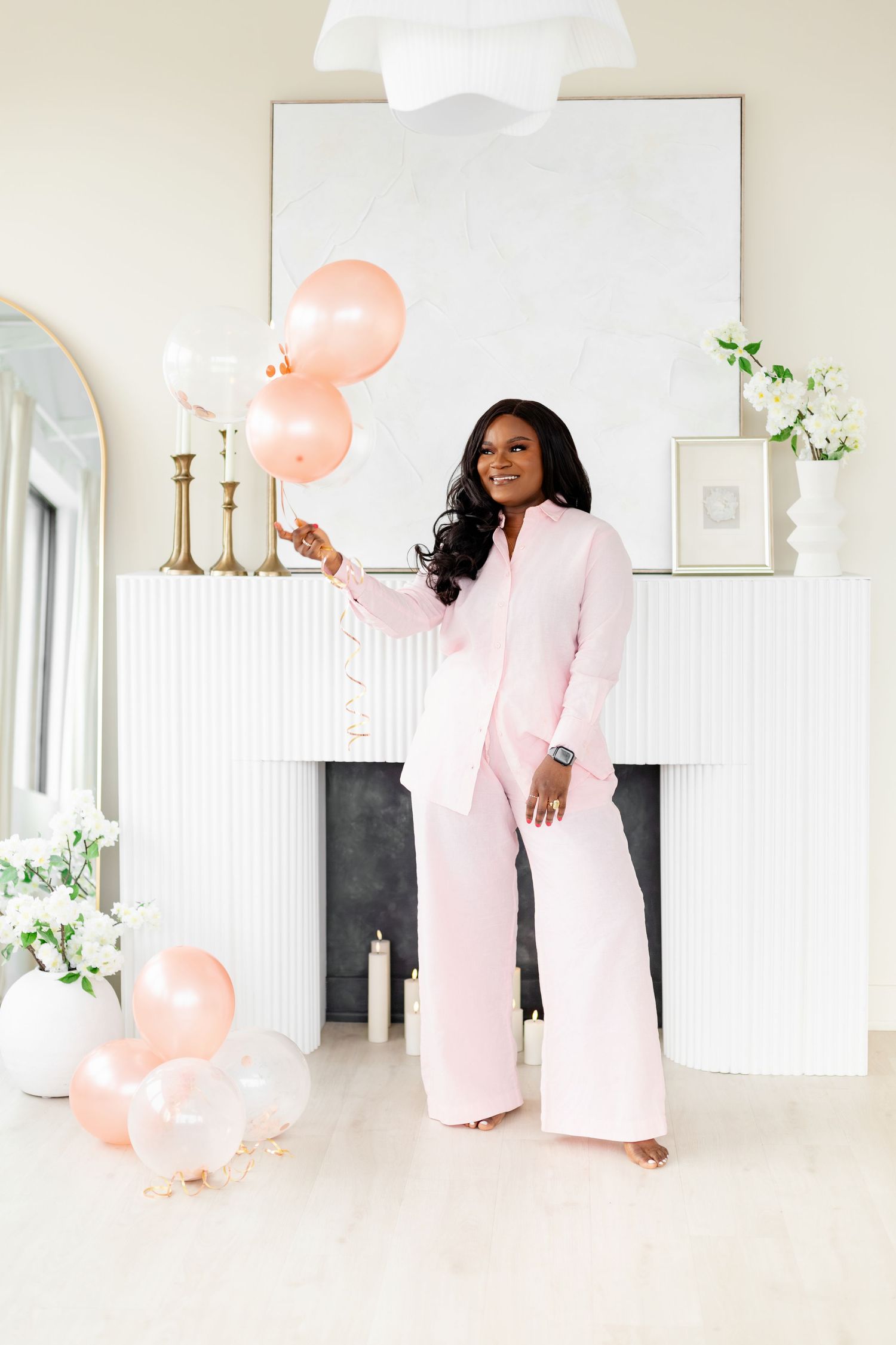 A figure in a flowing pink dress poses elegantly against a bright white interior with pink balloons and floral accents.
