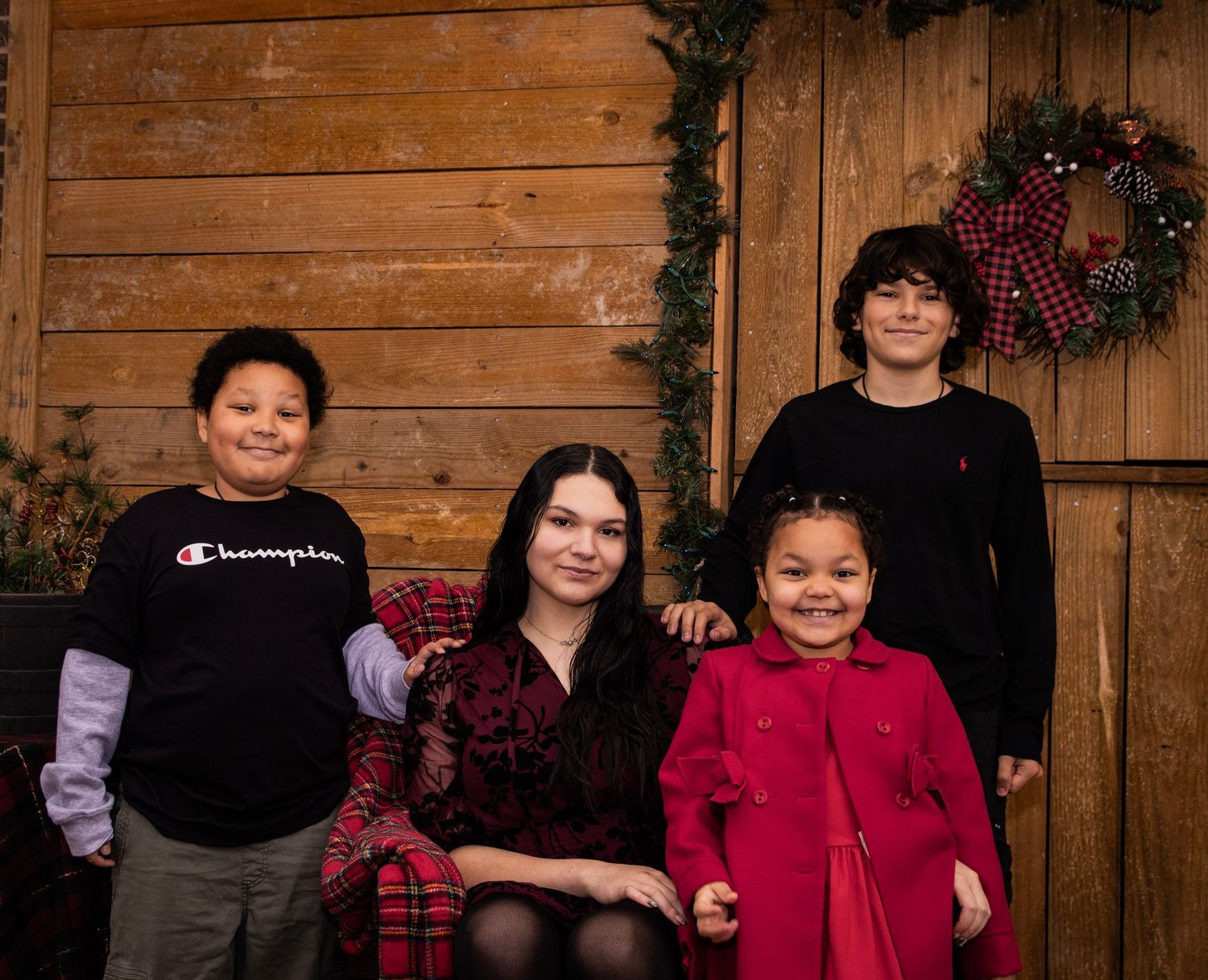 A family wearing coordinated red and black outfits poses together against a rustic wooden wall with holiday decorations.