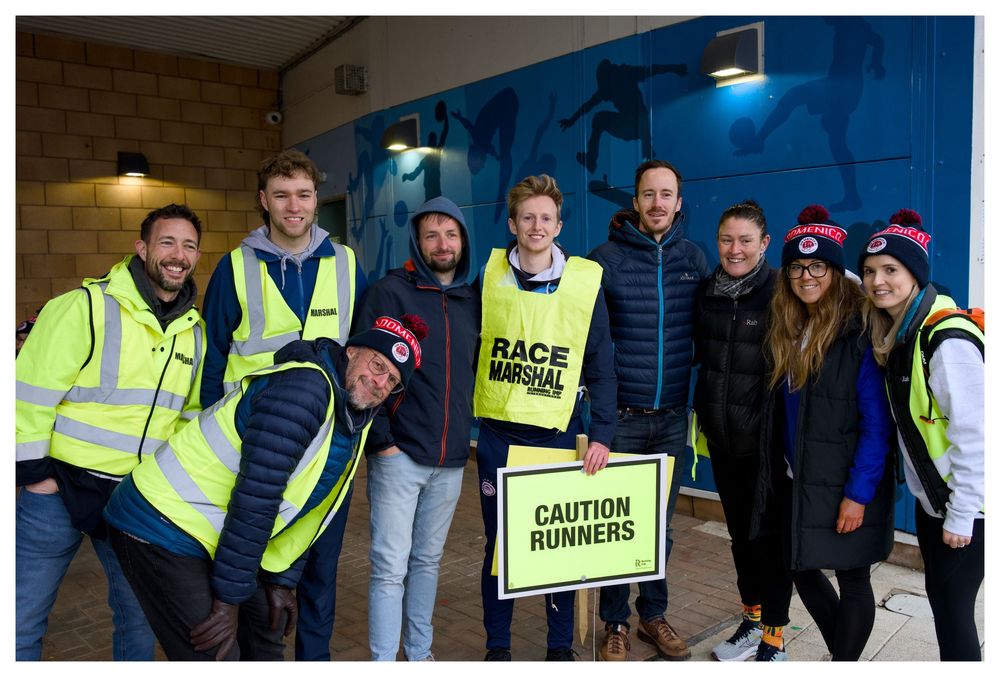 Group of race marshals posing with Caution Runners sign at outdoor running event wearing hi-vis vests and winter gear.