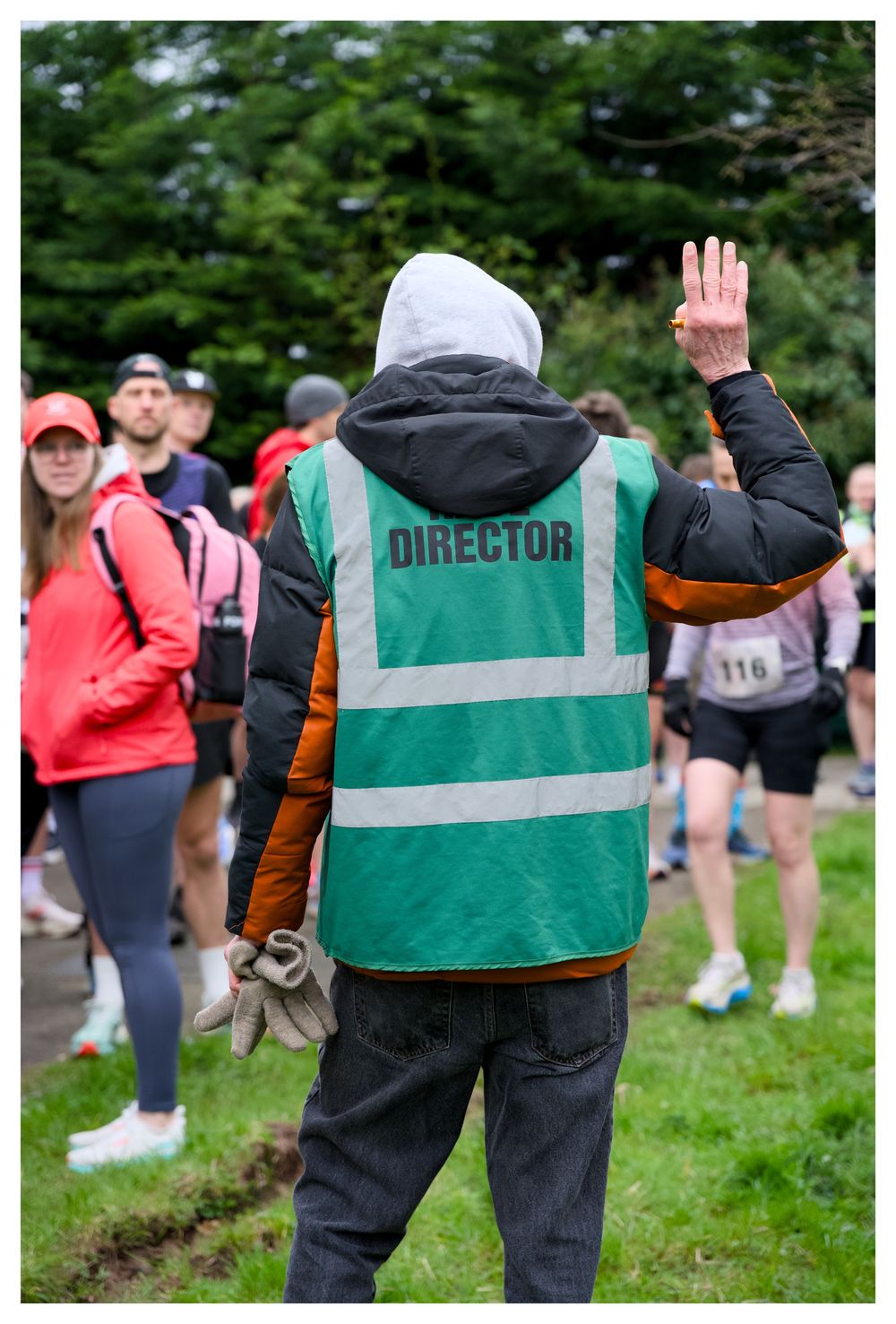 Race director in green high-visibility vest with DIRECTOR text waves to runners at outdoor running event.