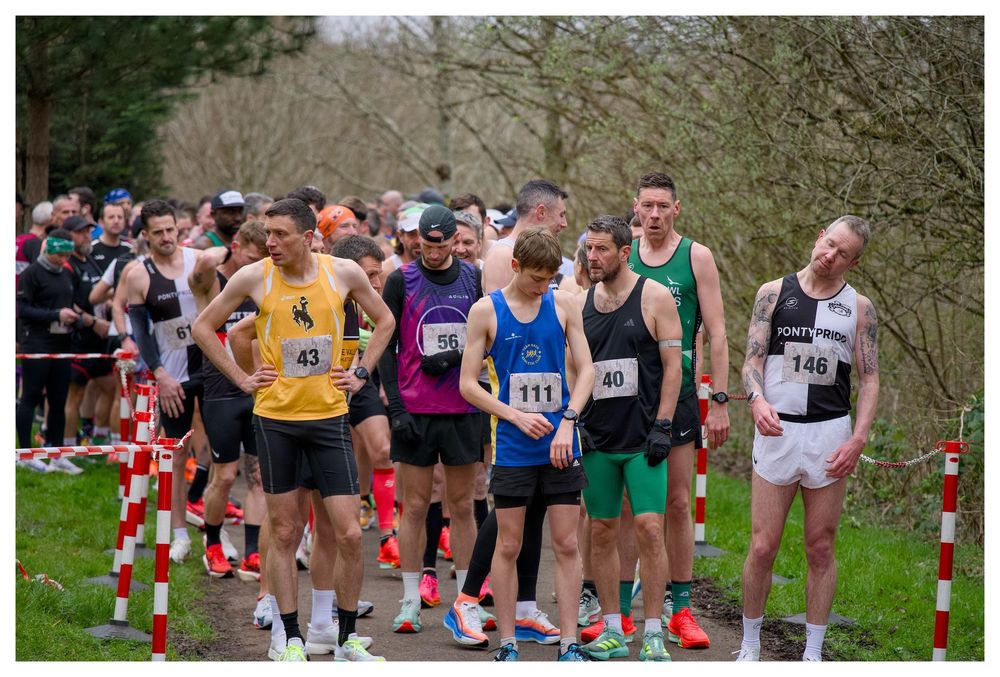 Male runners wearing numbered race bibs at the starting line of an outdoor cross country running race.