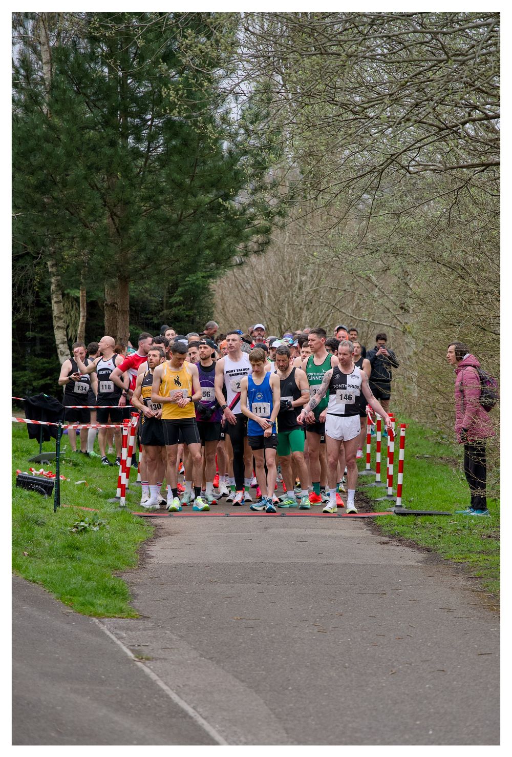 Runners gathered at the starting line of an outdoor road race in a tree-lined park on a cloudy day.