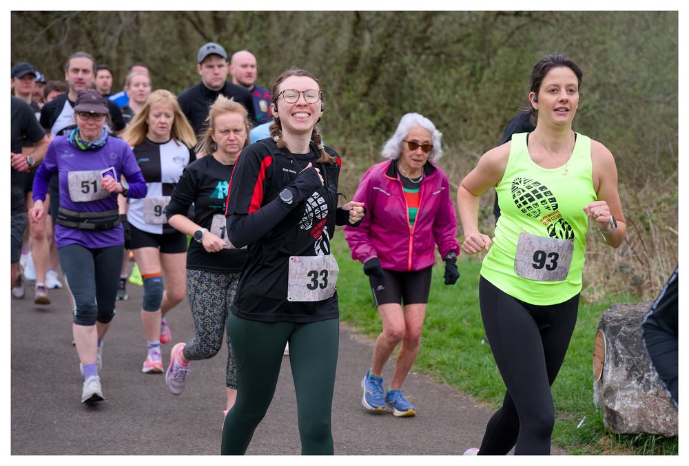 Female runners wearing race bibs 33 and 93 smile while competing in outdoor road race event.