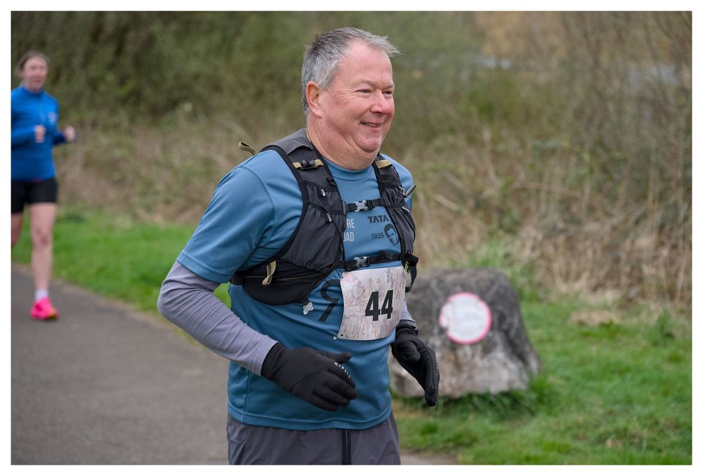 Smiling male runner wearing bib number 44 and hydration vest running on scenic outdoor race path.
