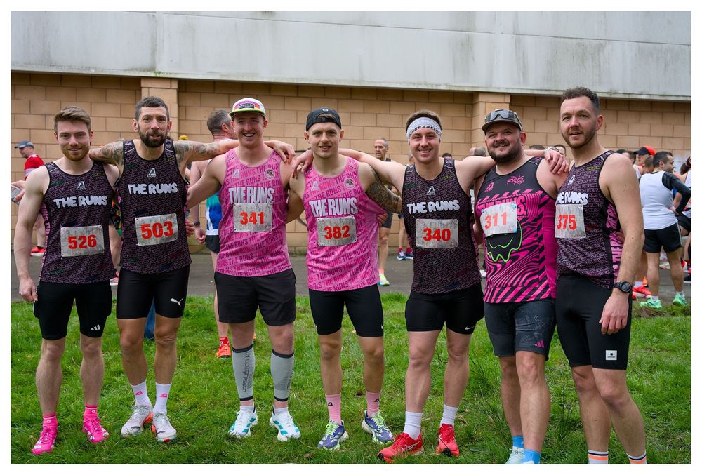 Group of seven male runners in matching pink and black The Duns team kit posing before a race.