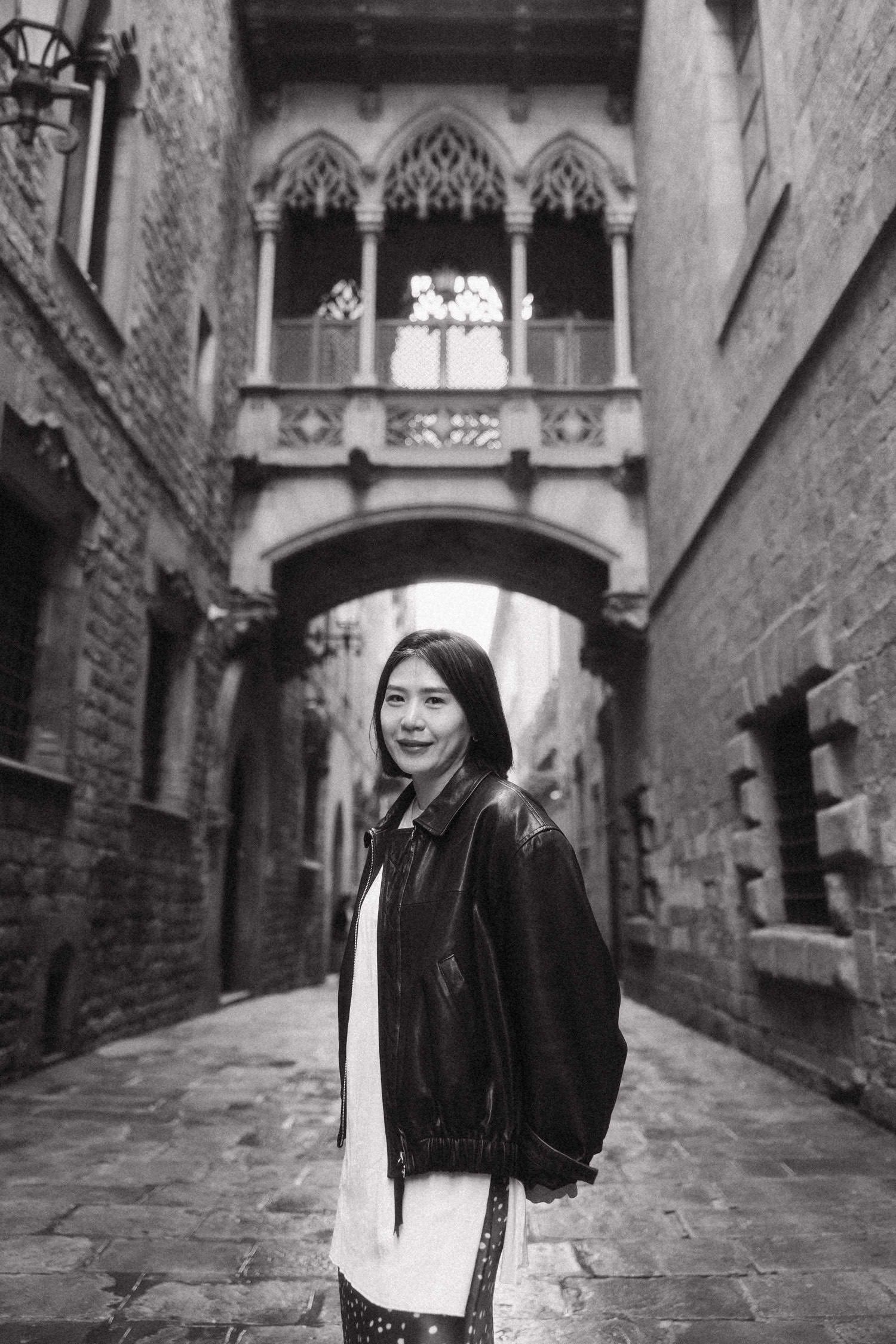 Black and white photo of a young Asian woman smiling in a historic Gothic alley with an ornate bridge overhead.