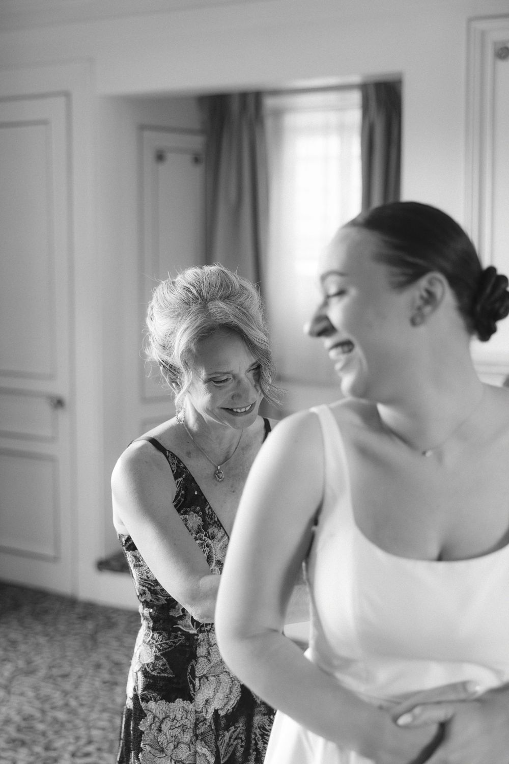 A black and white photograph showing joyful moments during wedding preparations in an elegant room with natural light.