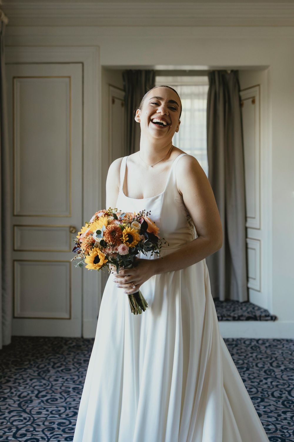 A joyful person wearing a white wedding dress holds a vibrant bouquet of orange and yellow flowers while laughing.