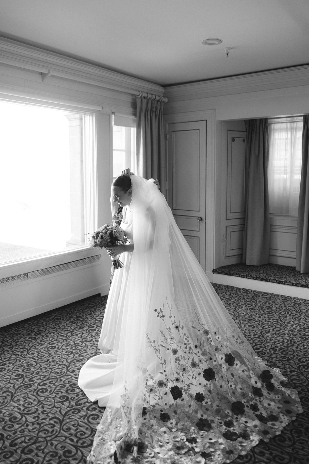 A flowing white wedding veil with delicate floral embellishments trails across a patterned hotel room carpet.