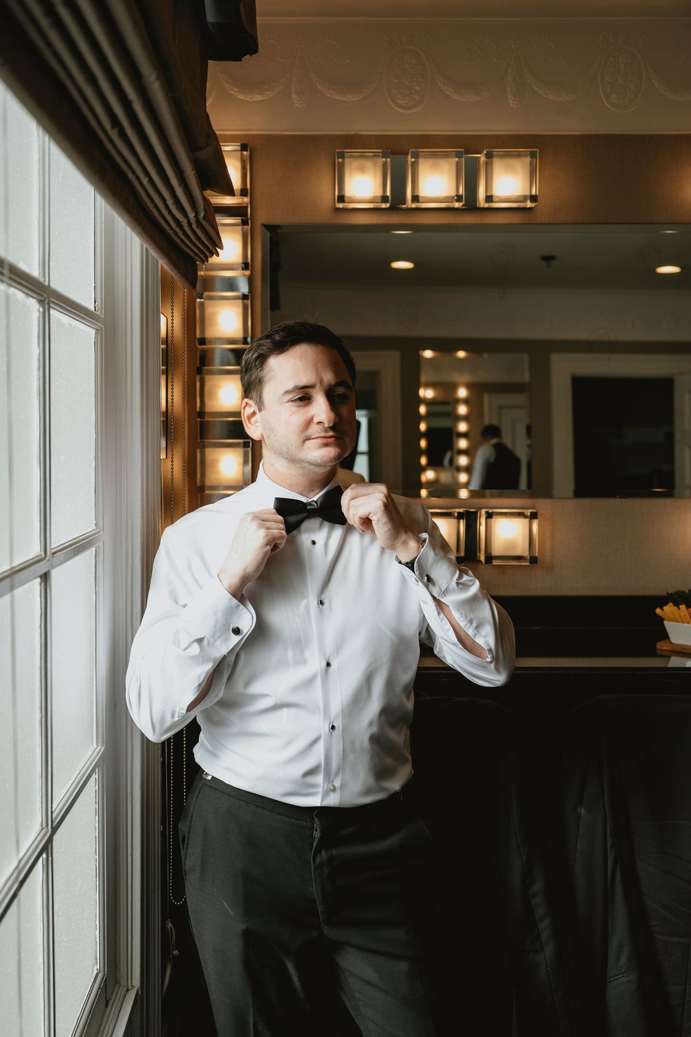 Someone adjusts their bowtie while standing by a window in formal attire at an elegant venue with warm lighting.