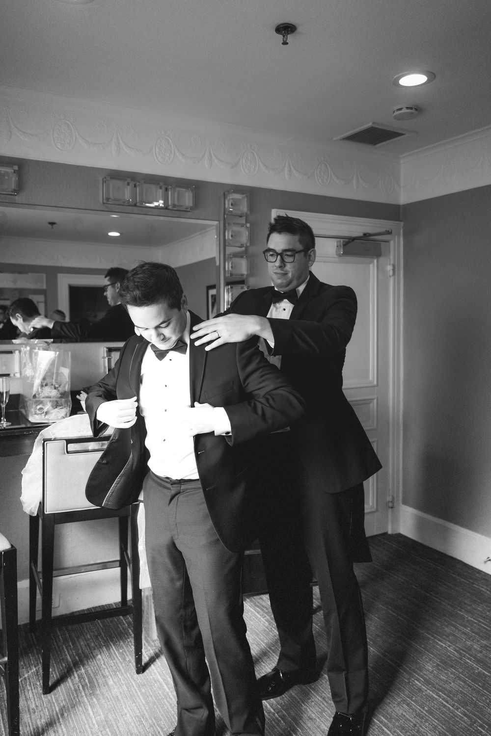 A series of black and white photos showing groomsmen helping adjust formal attire in a hotel room.