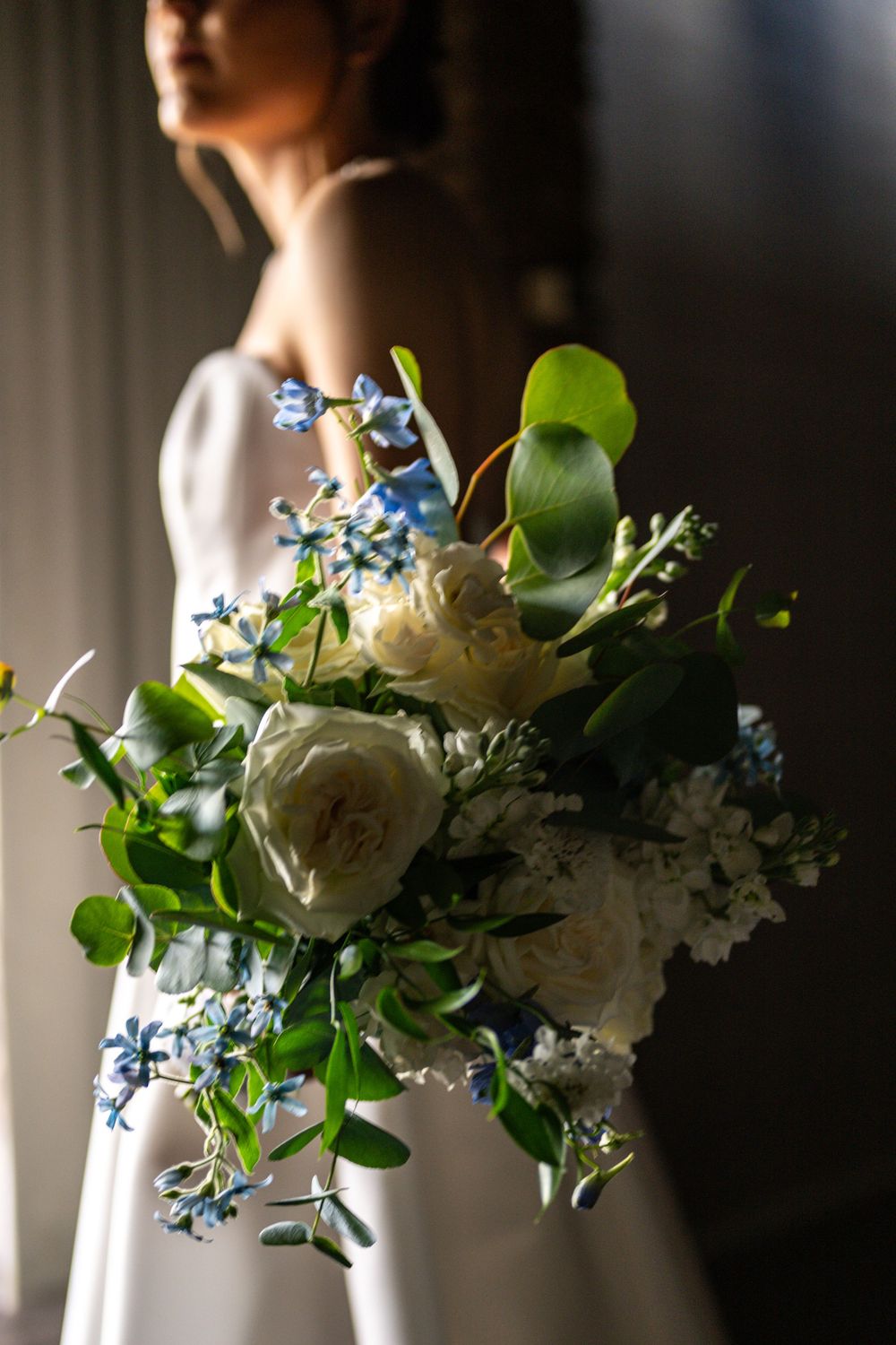 Bride in white dress holding a lush wedding bouquet with white roses, blue flowers, and green foliage in soft natural light.