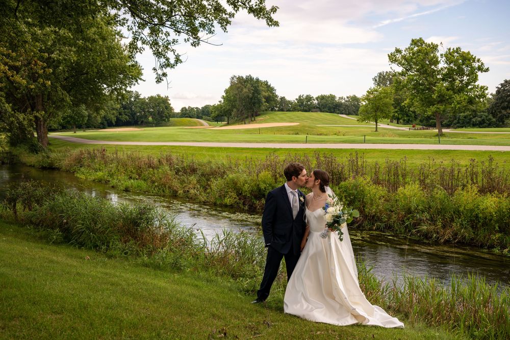 Bride and groom share a kiss beside a serene creek on a lush golf course on their wedding day.