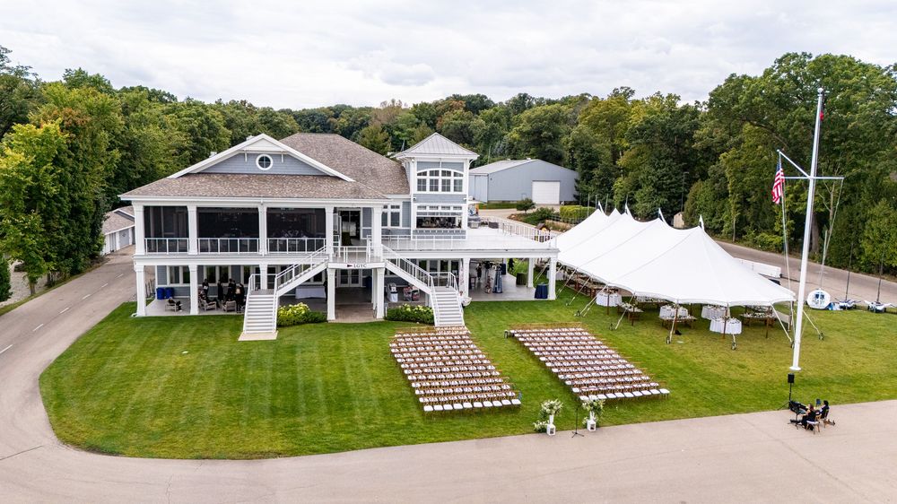 Aerial view of elegant white event venue with outdoor ceremony chairs and large tent on lush green lawn.