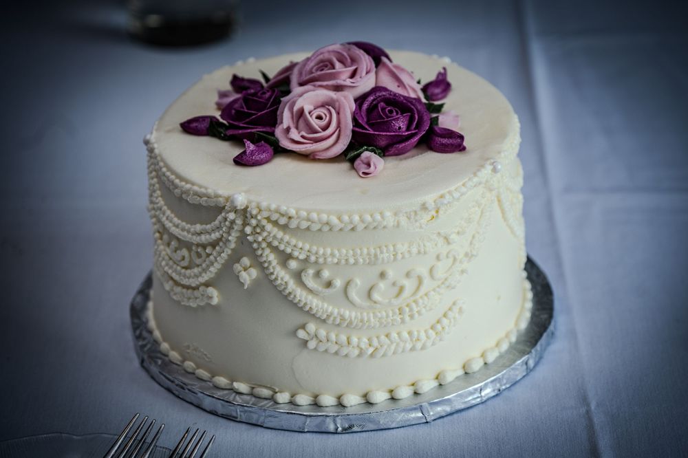 Elegant white wedding cake with purple and pink rose decorations and intricate pearl piping details on a silver cake board.
